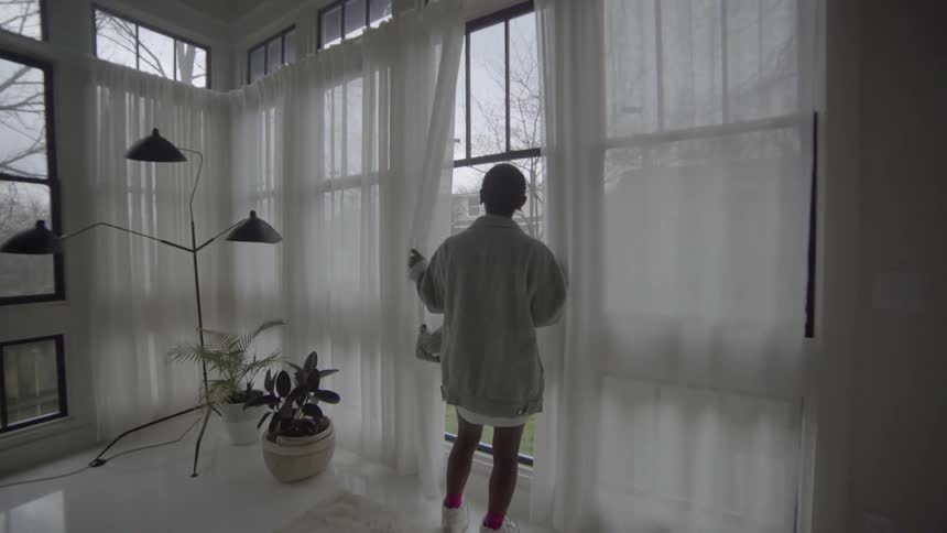 A woman moves the sheer drapes and looks out the window before walking away