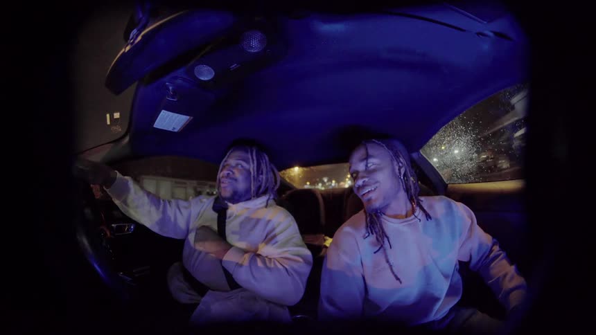 Two men shake hands and laugh and talk while in a car