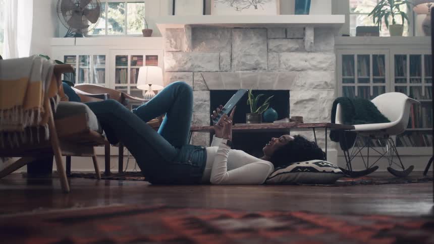 Black woman lying down on the floor uses a tablet in the living room at home