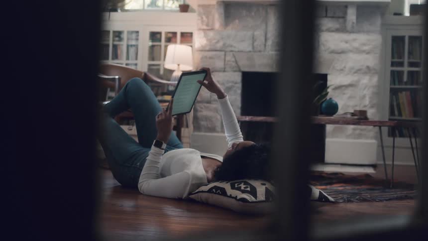 Black woman lying down on the floor uses a tablet in the living room at home