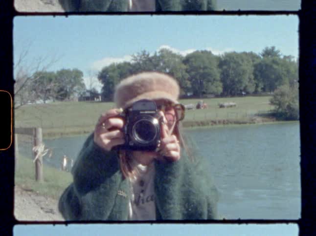 A woman with a camera taking a picture with a pond and a field in the background