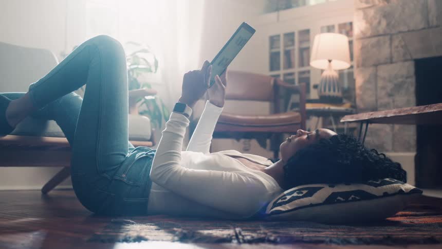 A woman laying on the floor with her head on a pillow and her feet on a couch while watching a tablet