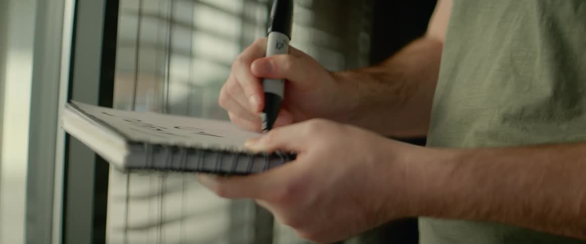 A man writing in a notebook with a marker while standing near a window