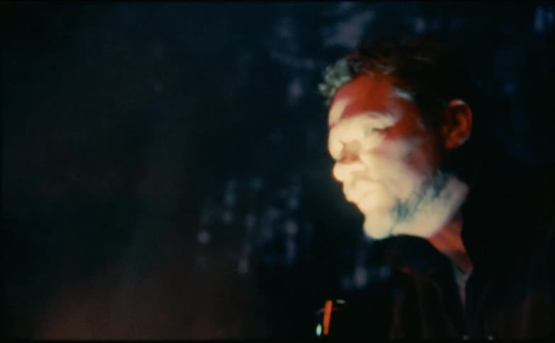 A man near a campfire shining a flashlight on his face while talking