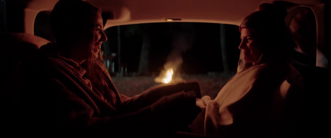 Two young women sitting inside a car close to a campfire talking and laughing together