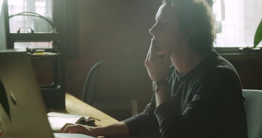 A man sitting at a desk rubs his chin as he thinks then goes back to his keyboard