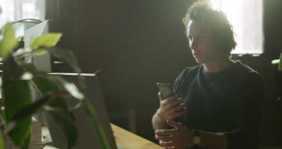 A man sitting in an office chairs turns and rolls over to his desk while holding his smart phone