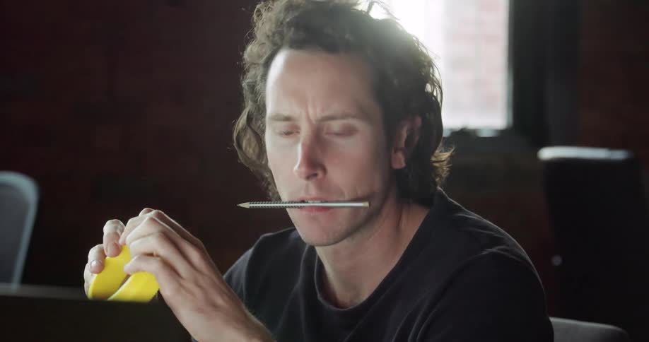 A man sits at a desk with a pencil in his mouth playing with a small yellow object and looking at a computer screen