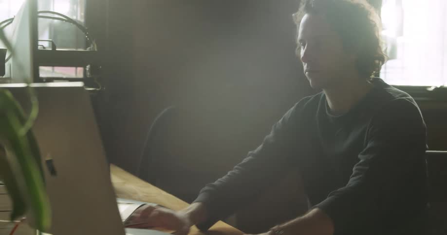 A man looking at a piece of plastic in a 3d printer then moving over to a laptop then typing