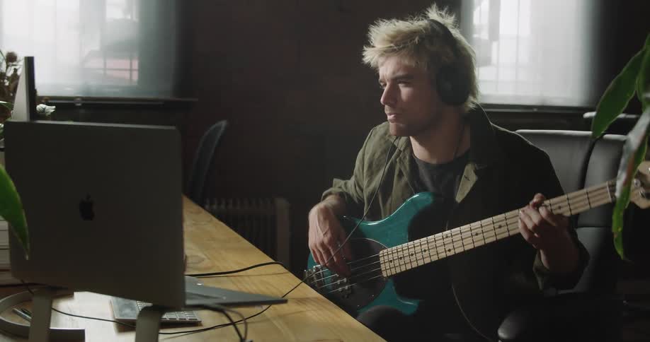 Man wearing headphones sits holding guitar in office studying monitor