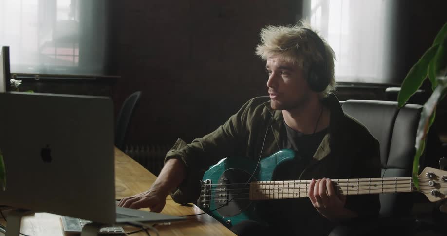 A man sits at a desk playing a guitar and working with a computer