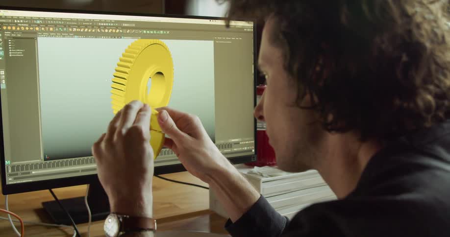 A man holds a yellow plastic ring turning it in his hands and comparing it to the yellow gear 3d image on his computer screen