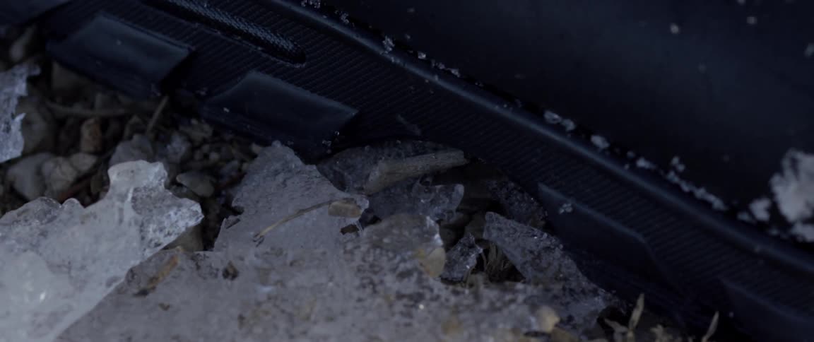 A layer of ice on the ground is crushed as someone wearing black boots walks on it