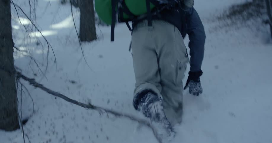 A man wearing a backpack struggles hiking up a snowy hillside in a forest