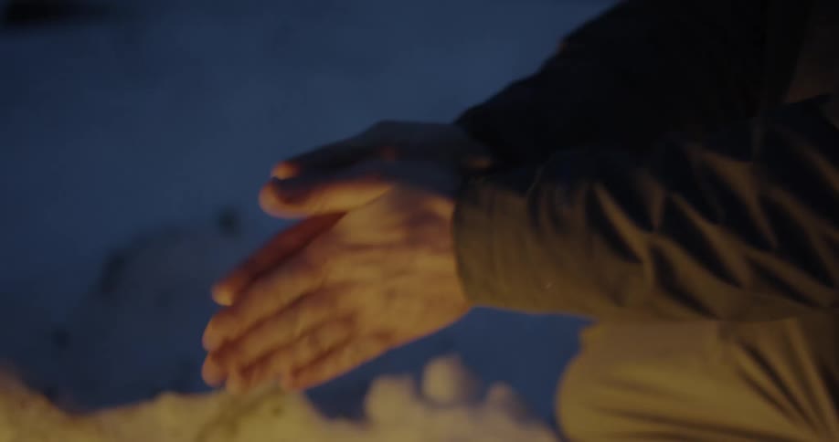 Close up of a person sitting near a camp fire in the snow warming up his hands