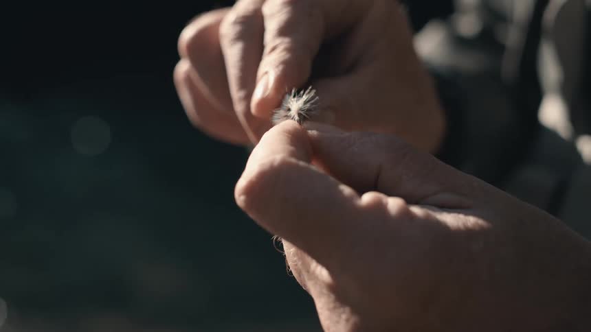 A man is grooming a fishing lure while outdoors near a lake