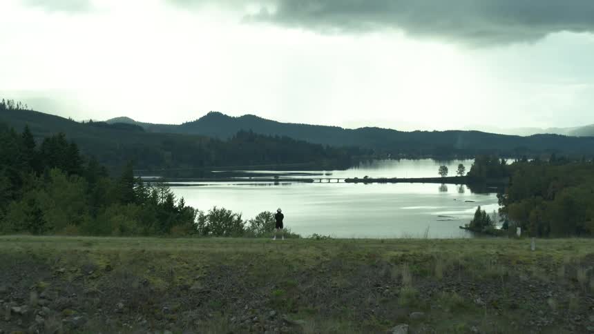 A man stands on a hill top looking over a pond and mountain scene