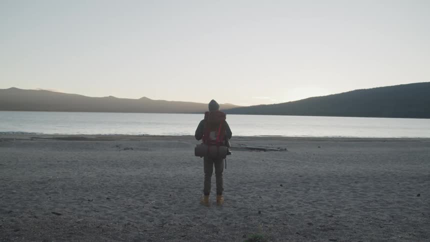 Person wearing hiking clothing and a backpack standing close to a lake and contemplating the view