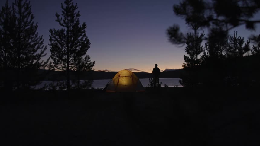 Mans silhouette standing next to the tent watches the lake at dusk