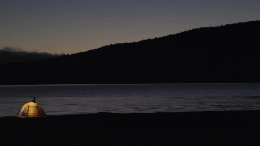 Man walking next to his yellow camping tent close to a lake at dusk