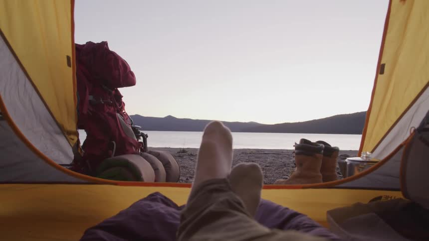 A persons feet with socks on relaxing in a tent with a lake in the foreground