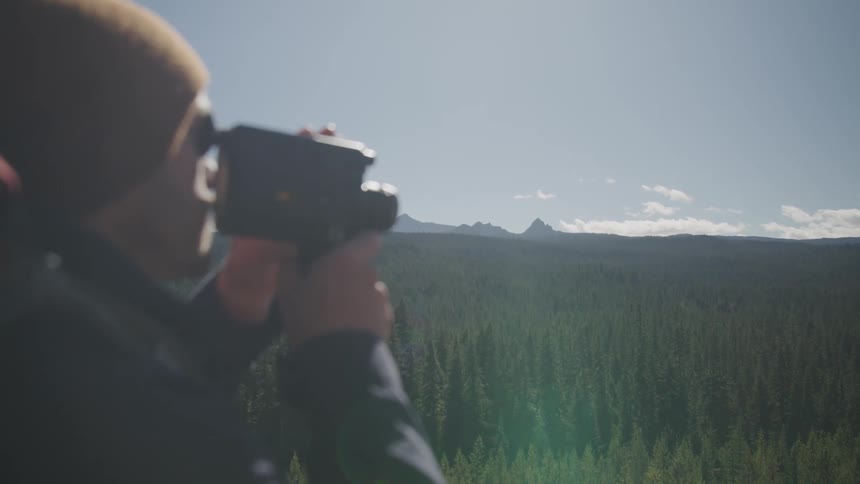 A man holds a camera to his face looking to a large forest area