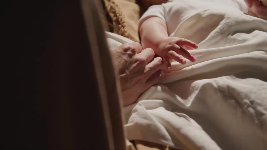 A baby reaches in his sleep while a parents hand sits on top of his white blanket