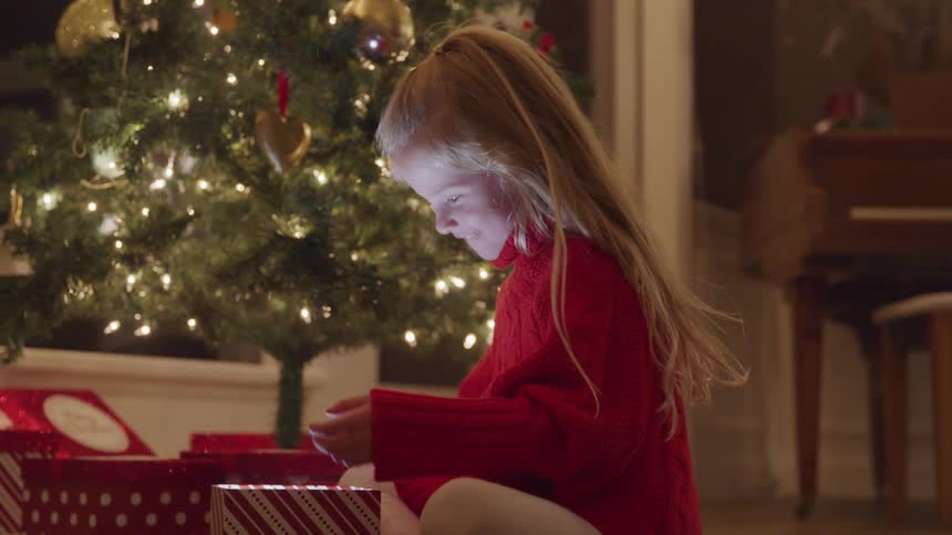 A young girl sits in front of a christmas tree and smiles as she opens a present