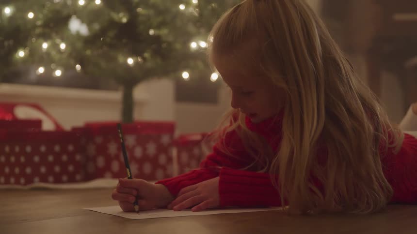 Little girl driwing on a paper while laying down in the ground next to a christmas tree