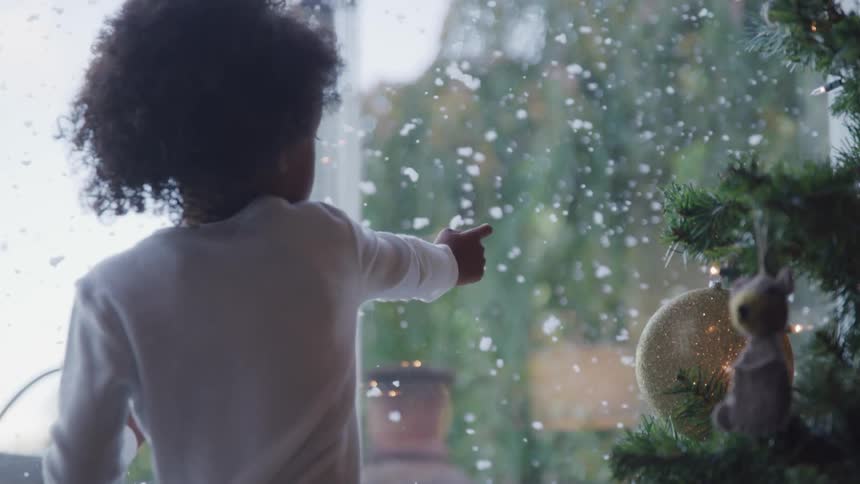 A child standing near his christmas tree looks out the large window and points at the falling snow