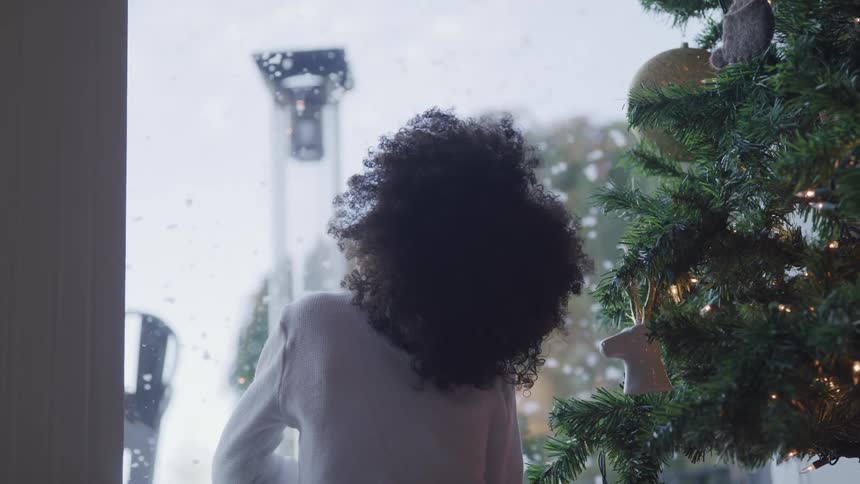 Black girl looking to the snow outside through the window next to a christmas tree