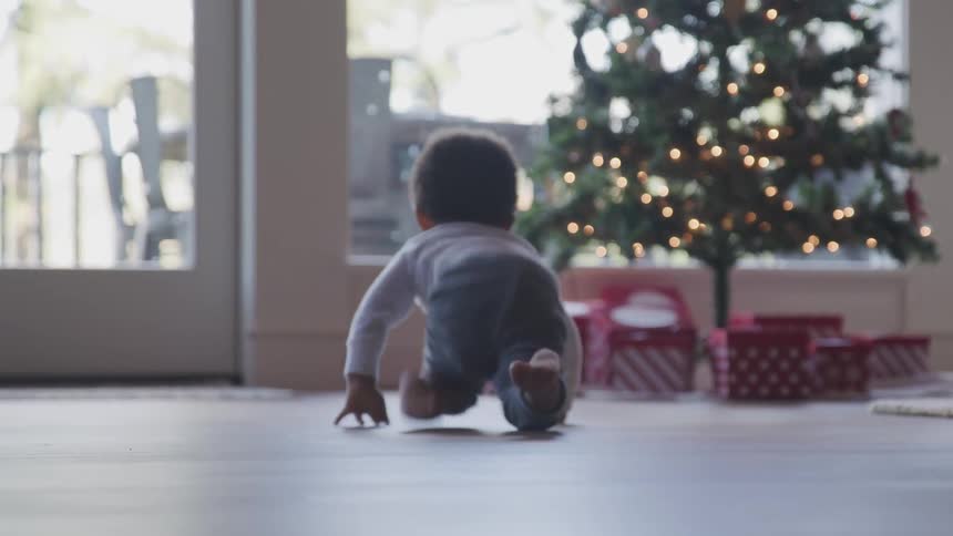 A baby crawls along the floor toward a christmas tree and presents