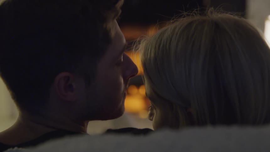 A man kisses his wifes forehead as they sit in front of a roaring fireplace