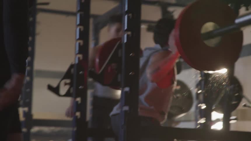 A man lifts heavy weight on his back in a gym while being cheered on by two others
