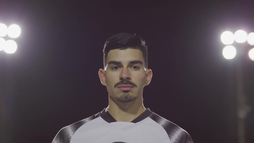 Male footballer stands and looks at the camera on the football field at night
