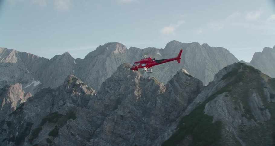 Red helicopter flying above mountains and clouds