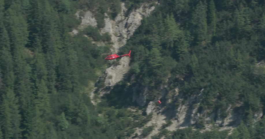 Red helicopter carries a basket as it flies over the forest