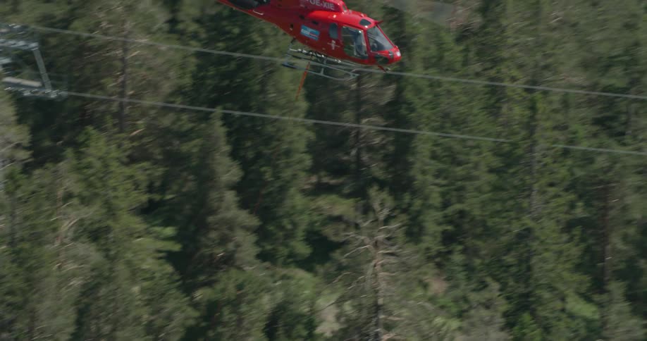 A helicopter using a cable to lift a large red container over a forest