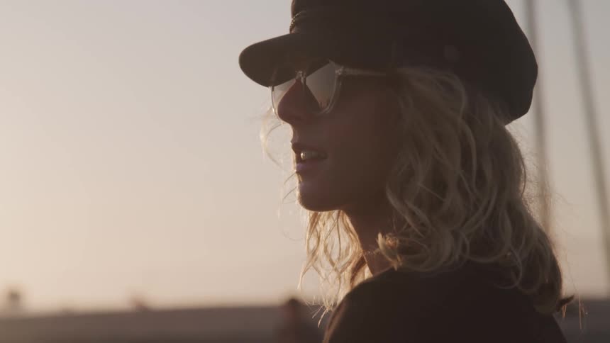 Woman wearing sunglasses and a hat looks around at sunset