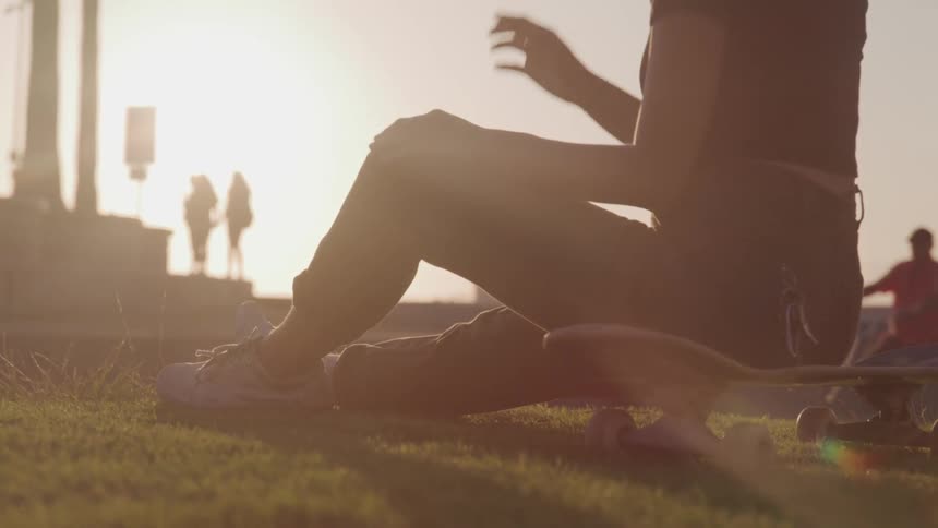 A woman sits on a skateboard on the grass at dusk while others are in the distance including one on a bicycle