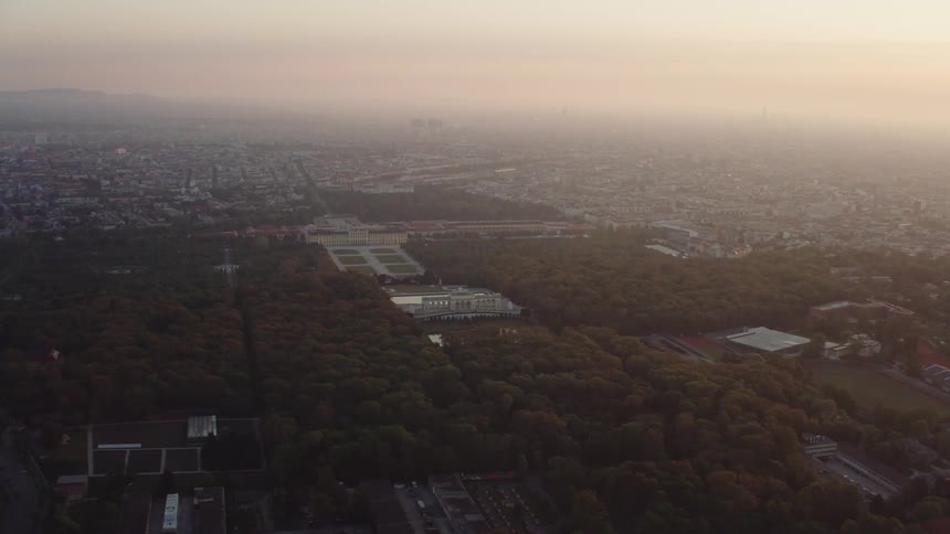 Ornate building complex and woods occupies center of sprawling town as sun tints fog on horizon pink