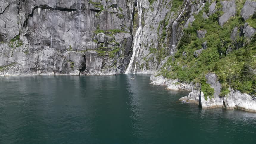 A cruise ship drives on an ocean with sailboats in front of a cliff that a waterfall is flowing into