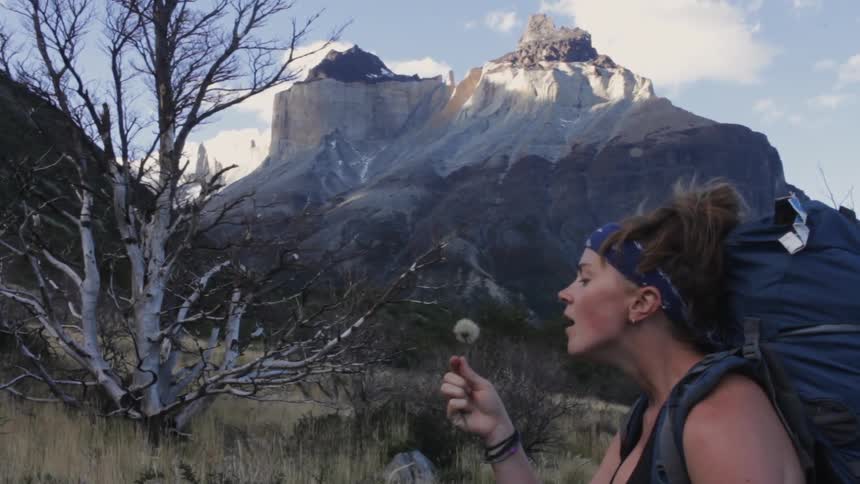 A woman is blowing a flower on a hillside