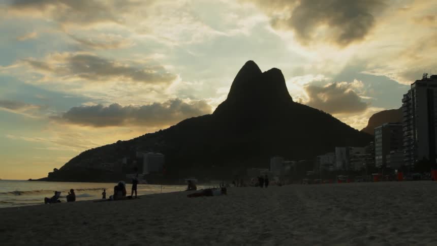People are near the beach as the sun sets behind a hill