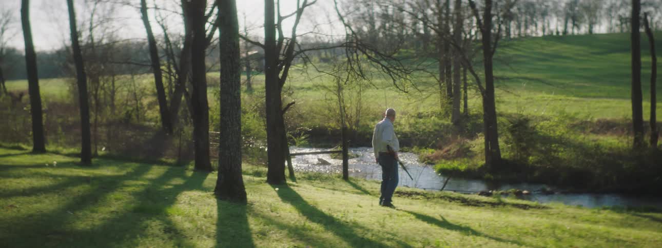 An elderly man carrying a shotgun approaches a stream and looks around