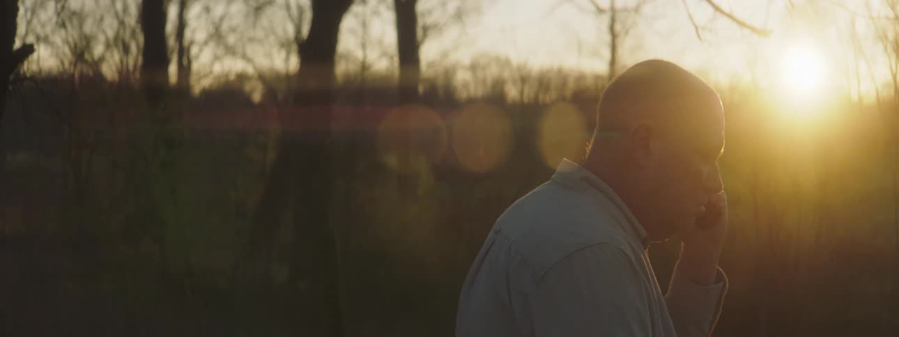 Older bald man stands among leafless trees talking on mobile phone as sun hovers near horizon