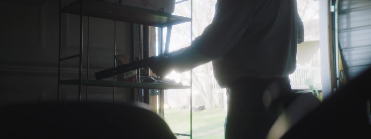 A mature man casually walks into a garage and retrieves a shotgun from a shelf