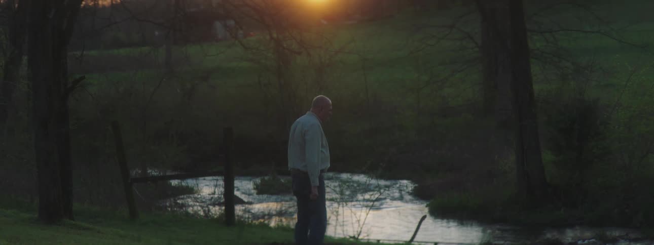 A man standing in front of a river drops a shotgun to the ground
