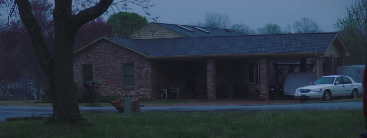 A white car is parked in the driveway of a ranch style home and other vehicles are under the carport covered with tarps
