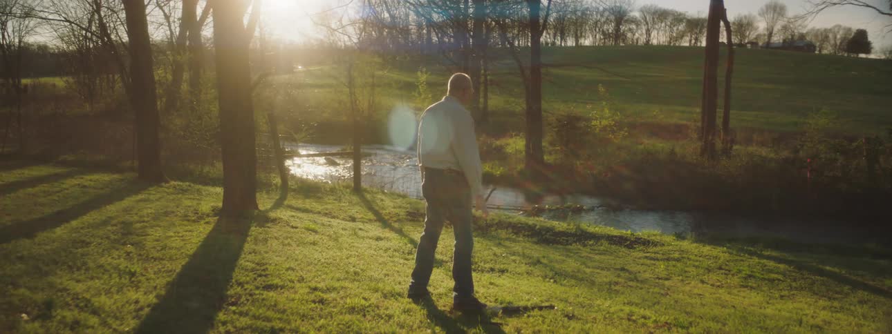 A man stands near a stream in the forest at dusk and suddenly drops his rifle